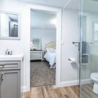 A modern bathroom with a view into an adjacent bedroom, featuring minimalist decor and a glass shower enclosure.