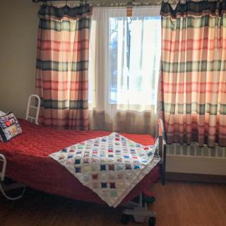 A small room with a hospital bed near a window dressed in patterned curtains and a colorful quilt on the bed.