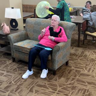 A senior woman enjoying a treat in a community lounge, surrounded by others engaging in various activities.