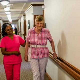 A healthcare worker assists an elderly woman walking down a corridor.