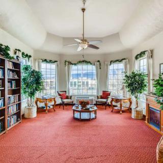 A cozy living room with large windows, bookshelves, and a seating area surrounded by plants.