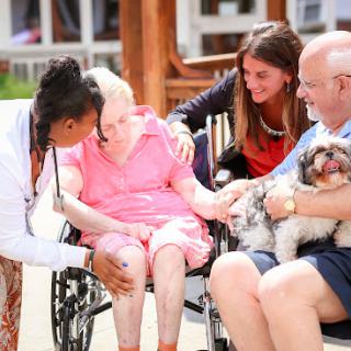 A caregiver and family members providing companionship and support to an elderly woman in a wheelchair, enjoying a sunny day with a pet.