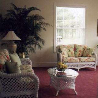 A cozy living room with wicker furniture and floral cushions, featuring a coffee table and an indoor plant next to a large window.