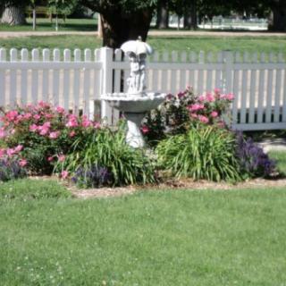 A charming garden with a small fountain surrounded by colorful flowers, bordered by a white picket fence.