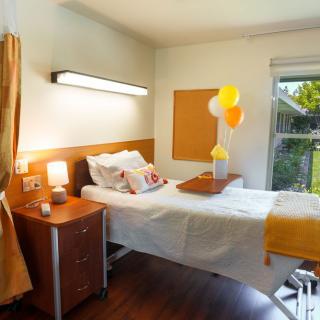 A bright hospital room with a neatly made bed, decorated with balloons and flowers, next to a window letting in sunlight.