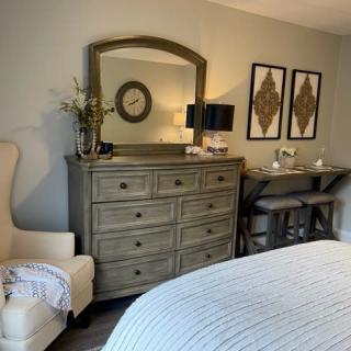 A cozy bedroom featuring a dresser with a mirror, decorated with a plant and framed artwork, alongside a comfortable chair and neatly made bed.