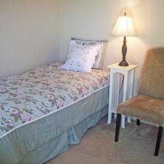 A cozy bedroom with a single bed, nightstand, and chair under a warm lamp.