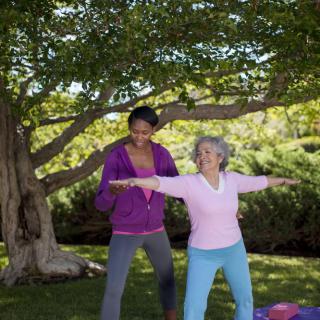 A yoga instructor guides an elderly woman in a yoga pose under a tree.