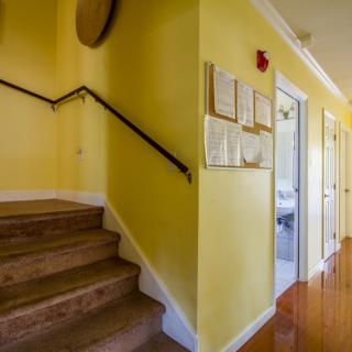 A brightly lit hallway with yellow walls and wooden floors featuring a staircase on the left.