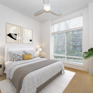 A modern and minimalist bedroom with white walls, featuring a neatly made bed, artwork on the wall, and a large window allowing natural light to fill the room.