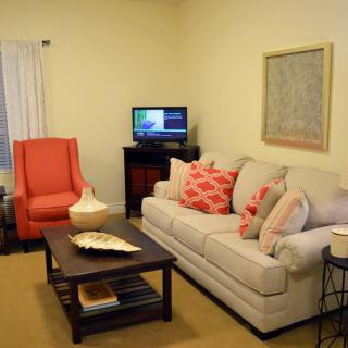 A cozy living room with a beige sofa, accented by red pillows and an armchair, centered around a wooden coffee table and a small television.