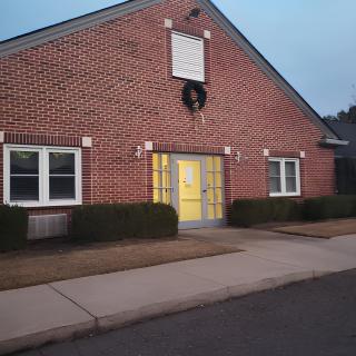 A brick building with a wreath above the yellow entrance door, flanked by windows and shrubs.