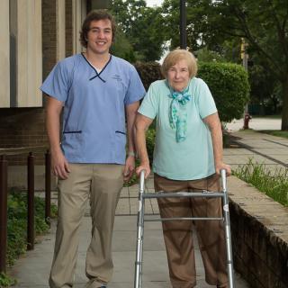 A caregiver assisting an elderly woman using a walker outside.