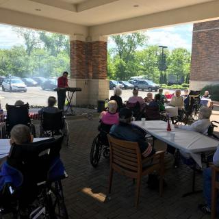 An outdoor musical performance attended by an audience, including elderly individuals in wheelchairs.