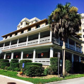 A large, multi-story building with a wraparound porch and a palm tree in front, set against a clear blue sky.