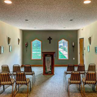 A small chapel interior with a pulpit and arranged seating facing a wall with windows and a cross.