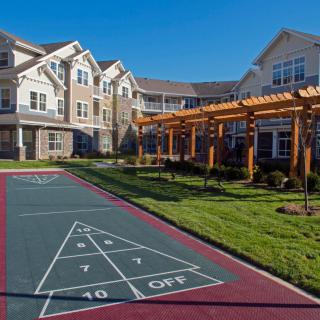 A sunny day view of a residential building with an outdoor shuffleboard court and a pergola.