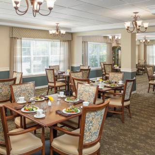 A well-lit dining area with elegantly set tables and chairs, featuring large windows and chandeliers.
