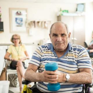 An elderly man holding a dumbbell while seated, with another person in the background, in an indoor setting.