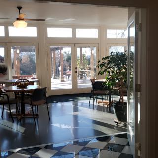 A well-lit dining area with large windows, tables, chairs, and a potted plant.