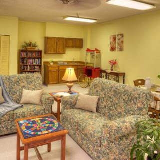 A cozy living room with floral-patterned sofas, a small table, and a lamp, adjacent to a kitchenette and bookshelf.