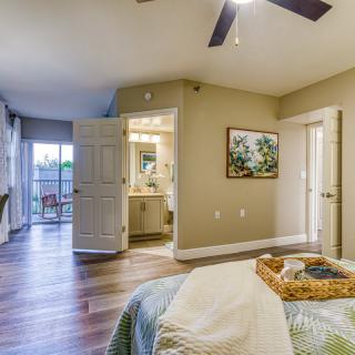 A cozy bedroom with a stylish interior, featuring a comfortable bed, ceiling fan, and wooden flooring. An open door leads to the bathroom, and artwork adorns the wall.