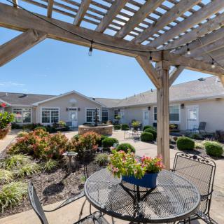 A sunny courtyard with a patio, pergola, and outdoor seating surrounded by well-maintained gardens.