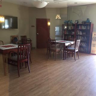 A cozy dining area featuring wooden tables and chairs, complemented by a bookshelf and warm lighting.