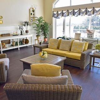 A well-lit living room featuring a wicker sofa with yellow cushions, matching armchairs, and a coffee table, complemented by decorative elements and large windows.
