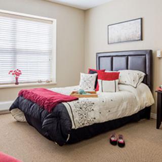 A well-decorated bedroom with a neatly made bed, adorned with red and white pillows and a matching throw blanket, brightened by natural light from the window.