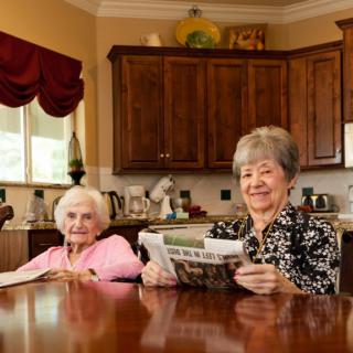 Two senior women sitting at a kitchen table, one is reading a newspaper and both are smiling.