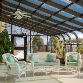 A bright and inviting sunroom featuring wicker furniture with green cushions, surrounded by lush plants and large windows.