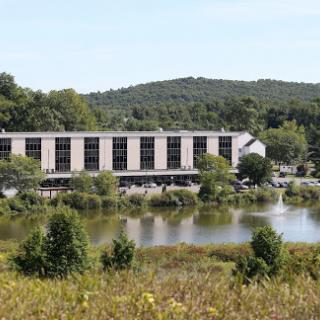 A serene view of a modern building by a tranquil lake, surrounded by lush greenery and hills.