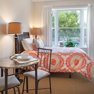 A modern bedroom with a colorful bedspread, a small dining table with two chairs, and large windows allowing natural light.