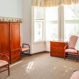 A bright room with wooden furniture including chairs, a dresser, and a large window.