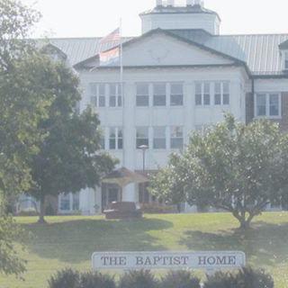 A large building with a sign reading 'The Baptist Home', surrounded by trees and an American flag.