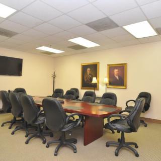 A conference room with a large meeting table surrounded by office chairs, decorated with portraits and a TV screen on the wall.