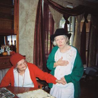 Two elderly women dressed in costumes, with one sitting and the other standing, sharing a light moment indoors.