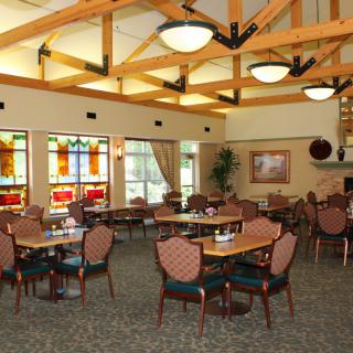 A spacious and well-lit restaurant interior with multiple tables and chairs, featuring decorative windows and overhead ceiling beams.