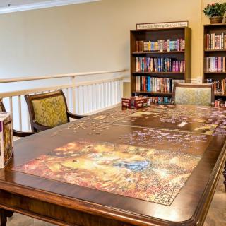 A cozy library setting featuring a wooden table ready for a board game session, surrounded by bookshelves filled with various books.