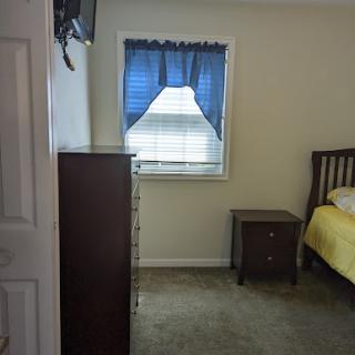 A cozy bedroom featuring a bed with yellow bedding, a dresser, and a nightstand beside a window with blue curtains.