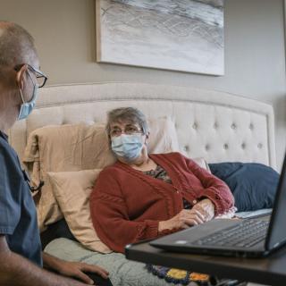 A healthcare worker visits a patient at home, both wearing masks, and engages in conversation with the help of a laptop.