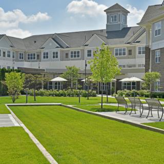 A well-maintained garden area in front of a large residential building with seating and umbrellas on a sunny day.