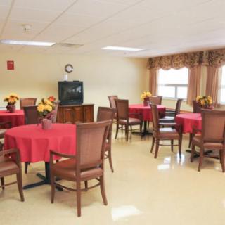 A dining room with several tables covered in red tablecloths and decorated with flowers, featuring a television and windows letting in natural light.