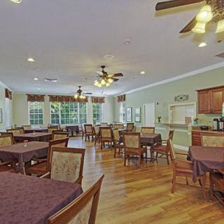 A spacious and bright dining room with multiple tables and chairs, featuring wooden flooring and ceiling fans.