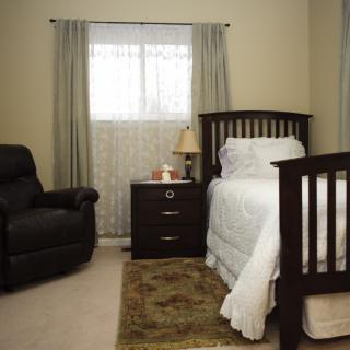 A cozy bedroom featuring a single bed, armchair, nightstand with a lamp, and window with curtains.
