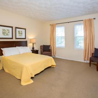 A well-decorated bedroom featuring a bed with a yellow cover, two chairs, decorative art on the wall, and windows with curtains.
