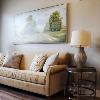 A cozy living room setting featuring a beige sofa adorned with decorative pillows, accompanied by an elegant lamp and a landscape painting.