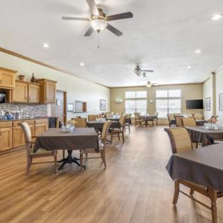 A spacious dining area with multiple tables and chairs, featuring a wood floor and ceiling fans, adjacent to a kitchen space.