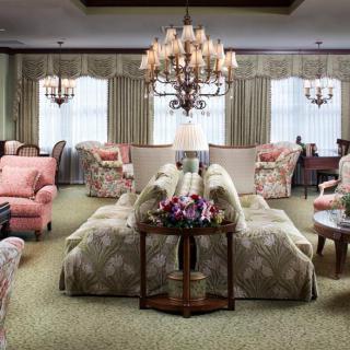 A traditionally styled living room with ornate chandeliers, floral patterned sofas and armchairs, and large windows with curtains.